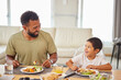 © peopleimages.com - Dad, kid and happy at table for lunch, healthy food and bonding in living room. Father, boy child and smile at home for nutrition, family time and Sunday meal with funny story or joking together