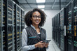 © polack - African American woman IT specialist with curly hair and glasses is smiling and holding a tablet. She is standing in a room full of servers