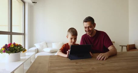 Cheerful positive dad helping little son to use application on tablet computer. Schoolkid aged boy and caring dad using digital pad at table home together, playing online game