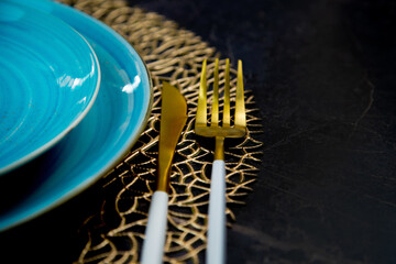 Wall Mural - Close-up. View of the part of the blue plates stand one on one on a gold patterned napkin, gold cutlery lies on the side: a fork and a knife on a black background. The concept of serving, the culture