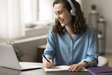 Canvas Print - Cheerful student girl listens audio course through wireless headphones, making notes, prepare assignment, sit at desk look at laptop screen, watch educational videos, take part in webinar. Tech, study