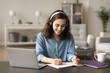 © fizkes - Beautiful smiling student girl makes assignment, listen audio course through headphones, learning subject on-line, sit at table with laptop, writes in copybook, prepare essay or exercise, do hometask