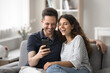 © fizkes - Beautiful married couple resting on sofa with smartphone, watching funny videos in social media platform, laughing, enjoy internet amusing content on weekend leisure. Fun, modern tech, pastime at home