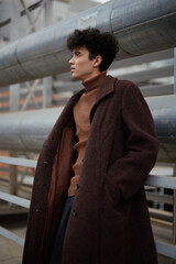 Wall Mural - Fashionable young man in a brown coat and turtleneck sweater, leaning against a metal railing with hands in pockets, gazing into the distance