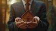 © AlexCaelus - A man in a suit holding a small wooden house model in a serene forest background, symbolizing real estate and home ownership.