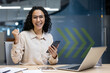 © Liubomir - Cheerful businesswoman celebrating success in modern office holding smartphone. Happy professional employee expressing achievement and joy while working on laptop. Concept of business victory