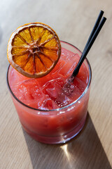 A glass of pink drink with a slice of orange on top. The drink is served in a short glass with a black straw