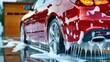 © WACHI - Red car in the process of being washed with soapy suds flowing down, reflecting sunlight near a wooden garage door.