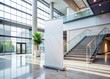 © Autun - Standing white roll-up banner template in modern business lobby, adjacent to sleek staircase, awaiting custom design, creating professional commercial display environment.