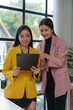© Phimwilai - Two confident Asian businesswomen stand holding tablets to discuss working on investment projects and strategic planning together at the office. brainstorming ideas.