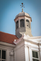 Close-up of Fatahillah Museum's iconic octagonal tower with a grey dome and golden weather vane. Featuring red tile roof, white colonial-style walls, and green shuttered windows against a clear blue s