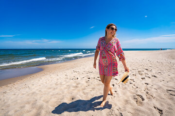 Beach holidays. Relaxation and sightseeing. Young woman wearing fashionable multicolored pink dress walking and holding beige hat in her hand on sandy beach on sunny day. Front view