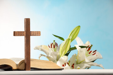 Wall Mural - Wooden cross, Bible and beautiful lily flowers on white table against light blue background, closeup. Religion of Christianity