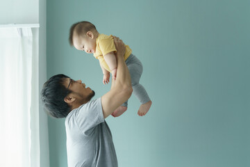 Asian Father lifting baby in yellow shirt and grey leggings in the air against blue wall, both smiling, Candid family moment, love and bonding concept.