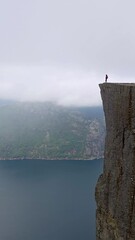 Wall Mural - A woman is standing on a cliff, admiring a tranquil lake amidst a picturesque landscape. The camera beautifully captures the serenity and isolation of this moment, Preikestolen Norway