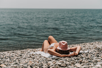 Canvas Print - A woman is laying on the beach with a pink hat on her head. She is wearing a white bikini and is laying on a towel. The beach is rocky and the water is calm. Scene is relaxed and peaceful.