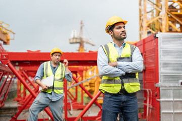 Wall Mural - Confident male engineer wearing a yellow hard hat and safety vest standing with arms crossed in front of tower crane equipment at construction site.