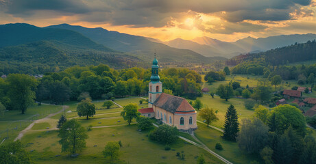 Wall Mural - Aerial view of a small church in the middle of a beautiful green valley, with forests on surrounding mountains