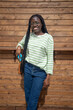 © wifesun - Smiling African American woman in glasses and striped shirt poses confidently against a wooden wall, enjoying a sunny day. Black young girl with a phone looks at the camera and smiles.