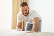 © Prostock-studio - A man in a white shirt and grey pants smiles while making his bed in his bedroom. He is kneeling on the bed, smoothing out the sheets with one hand. Sunlight streams through a window behind him.