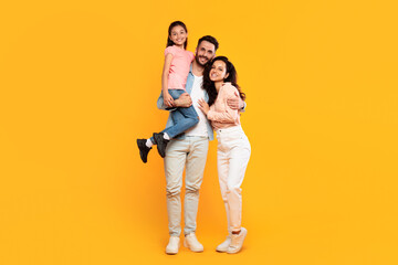 Wall Mural - A smiling family stands together against a bright yellow background. The father holds his daughter in his arms, while the mother stands beside him, her arm around his waist