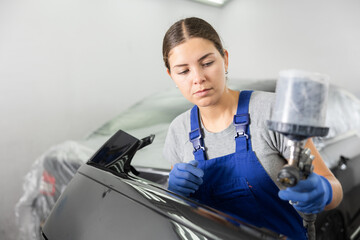 Wall Mural - Professional young female auto painter in blue overalls working in automotive workshop, focused on painting part of black car body using spray gun