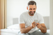 © Prostock-studio - A man in a white shirt sits on a white bed, smiling as he looks at his smartphone. He is sitting with his legs crossed and his arms are resting on his knees. The bed is made with a white sheet.
