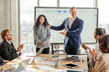 A group of business professionals gathered around a conference table in a modern office space are applauding a young lady colleague who has just finished giving a presentation