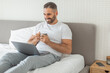 © Prostock-studio - A man is sitting in bed, working on a laptop computer while smiling and drinking from a mug. He is wearing a white t-shirt and gray sweatpants. A smartphone is on a nightstand next to the bed.
