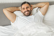 © Prostock-studio - A man with a beard and short hair is lying in bed, smiling and looking directly at the camera. He is wearing a white t-shirt and his hands are behind his head, resting on a white pillow.