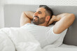 © Prostock-studio - A man with a beard lies in bed with his hands behind his head, looking off to the side with a relaxed expression on his face. He is wearing a white t-shirt and is covered by a white duvet.