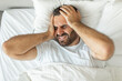 © Prostock-studio - A man lies in bed, his face contorted in pain as he holds his head with both hands. He is likely suffering from a headache or migraine, his expression showing discomfort and distress, top view