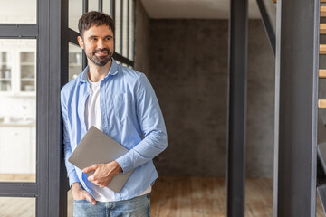 A young man with a beard is leaning against a glass and metal doorway while holding a closed laptop computer. He is dressed in a blue button-down shirt over a white t-shirt and blue jeans, copy space