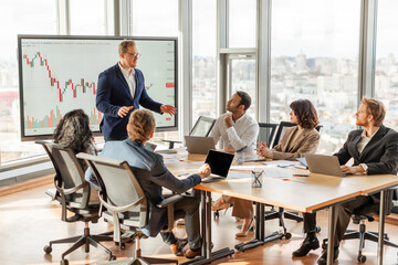 Aufkleber - A businessman is presenting financial data to a team in a modern office with large windows overlooking the city. He is standing and pointing at a chart on a screen