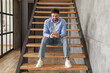© Prostock-studio - A man sits on a set of wooden stairs in a modern home. He wears a blue shirt, white t-shirt, blue jeans, and white sneakers. He is looking down at his phone and smiling.