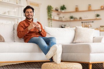 Wall Mural - Arab man is sitting on a white couch, his legs resting on a small wooden ottoman. He is wearing a light brown shirt, blue jeans, and white sneakers. He is smiling and looking down at his phone.