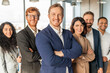 © Prostock-studio - A group of diverse business professionals stands in an office setting, all smiling confidently and looking at the camera. They are wearing professional attire, man in the front has his arms crossed