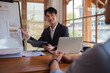 © wattana - businessman is conducting a presentation in a modern office setting. He is seated at a wooden table with a laptop and documents.