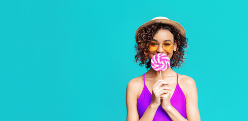 Wall Mural - A young woman with curly hair, wearing a purple swimsuit, sunglasses, and a straw hat, smiles while holding a pink and white lollipop in front of her face. She stands against turquoise background.