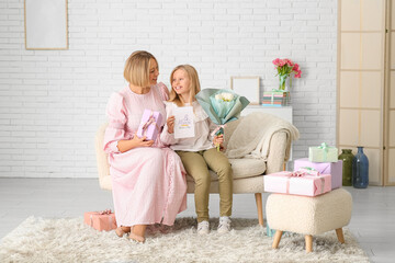 Poster - Beautiful young woman holding gift box and her cute little daughter with bouquet of white tulips and greeting card on sofa in living room