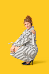 Wall Mural - Beautiful young woman in stylish striped dress sitting against yellow background