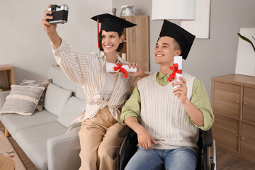 Wall Mural - Male graduate in wheelchair and his classmate with diplomas taking selfie at home