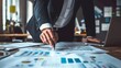 © piyapiya - A businessman in a suit points at financial documents spread across his desk.