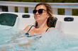© Lazy_Bear - Woman wearing sunglasses and swimsuit relaxing in outdoor hot tub with bubbling water and enjoying sunny day. Female resting in outdoor jacuzzi with beautiful natural landscape