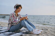© ViDi Studio - Full body young woman wear shirt casual clothes sits listen to music in headphones use mobile cell phone rest on sea ocean sand shore beach outdoor seaside in summer day free time. Lifestyle concept.