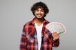 © ViDi Studio - Young happy cheerful smiling Indian man he wears red shirt casual clothes hold in hand fan of cash money in dollar banknotes isolated on plain grey color background studio portrait. Lifestyle concept.