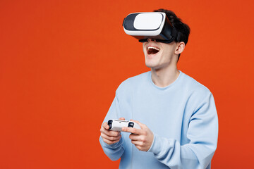 Wall Mural - Young man he wearing blue sweatshirt casual clothes hold in hand play pc game with joystick console watching in vr headset pc gadget isolated on plain red orange color background. Lifestyle concept.