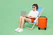 © ViDi Studio - Full body traveler IT woman wear casual clothes sit in deckchair hold use laptop pc computer work isolated on plain green background. Tourist travel abroad in free time rest. Air flight trip concept.