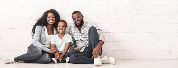 Wall Mural - Happy Millennial Family Portrait. Joyful Black Parents Posing Together With Their Little Daughter, Sitting On Floor Over White Brick Wall Background.