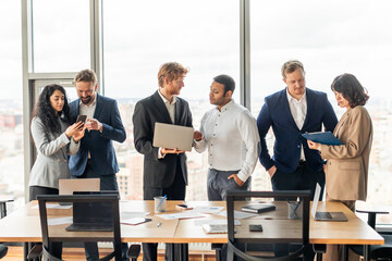 Aufkleber - A group of business professionals stand in a modern office space, discussing business while looking at a laptop and a clipboard. The office has large windows with a city view in the background